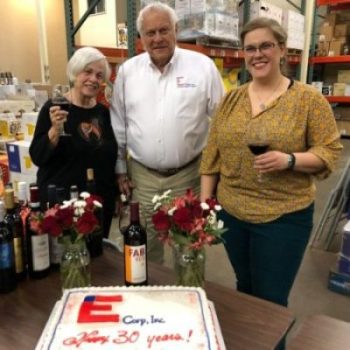 Alsn Carol and Dale with Cake 30th anniversary Carol Ann, Dale, and Allison Elliott standing behind a sheet cake with the E-Corp logo, celebrating their 30th anniversary.