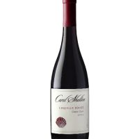 A burgundy-style dark glass wine bottle with a red matte capsule and a white label that says Carol Shelton Coquille Rouge in black calligraphy, with a small red stylized seashell at the bottom left corner.